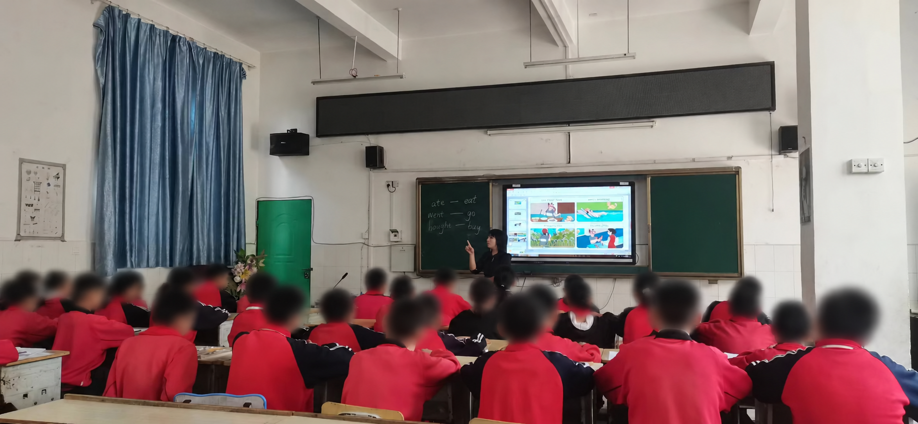 Students seated in an English classroom facing a teacher and a digital whiteboard displaying teaching materials. Student heads and faces are blurred in this edited copy.